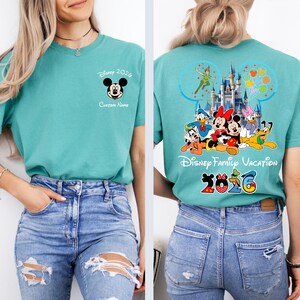 Op de afbeelding: Tealkleurig T-shirt met Mickey Mouse en de tekst "Disney 2026" op de voorkant. De achterkant toont Disney-personages, een kasteel en "Disney Family Vacation 2026".