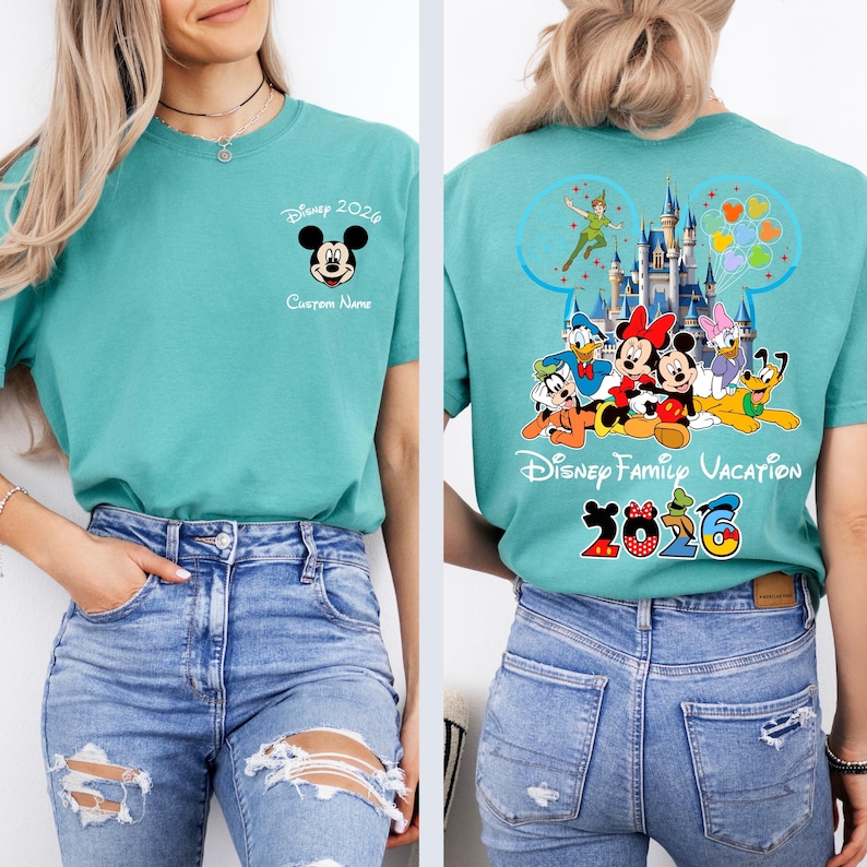 Op de afbeelding: Tealkleurig T-shirt met Mickey Mouse en de tekst "Disney 2026" op de voorkant. De achterkant toont Disney-personages, een kasteel en "Disney Family Vacation 2026".