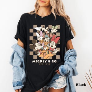 May include: Black t-shirt featuring a colorful graphic of Mickey Mouse, Minnie Mouse, Donald Duck, Daisy Duck, Goofy, and Pluto. The design is set against a checkered background with the text "Mickey & Co. Est. 1928".