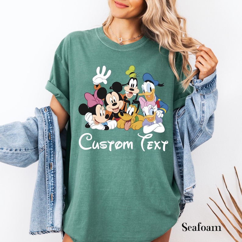 May include: Seafoam green t-shirt with a graphic of Disney characters: Mickey Mouse, Minnie Mouse, Donald Duck, Daisy Duck, Goofy, and Pluto. The words "Custom Text" are written in white script across the bottom of the graphic.