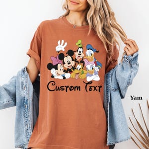 May include: A terracotta-coloured t-shirt with a graphic of Disney characters, including Mickey and Minnie Mouse, Goofy, Donald and Daisy Duck, and Pluto. The words "Custom Text" are written in a script font below the characters. The shirt has rolled-up sleeves.