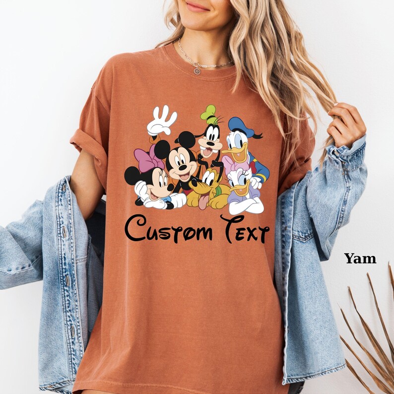 May include: A terracotta-coloured t-shirt with a graphic of Disney characters, including Mickey and Minnie Mouse, Goofy, Donald and Daisy Duck, and Pluto. The words "Custom Text" are written in a script font below the characters. The shirt has rolled-up sleeves.