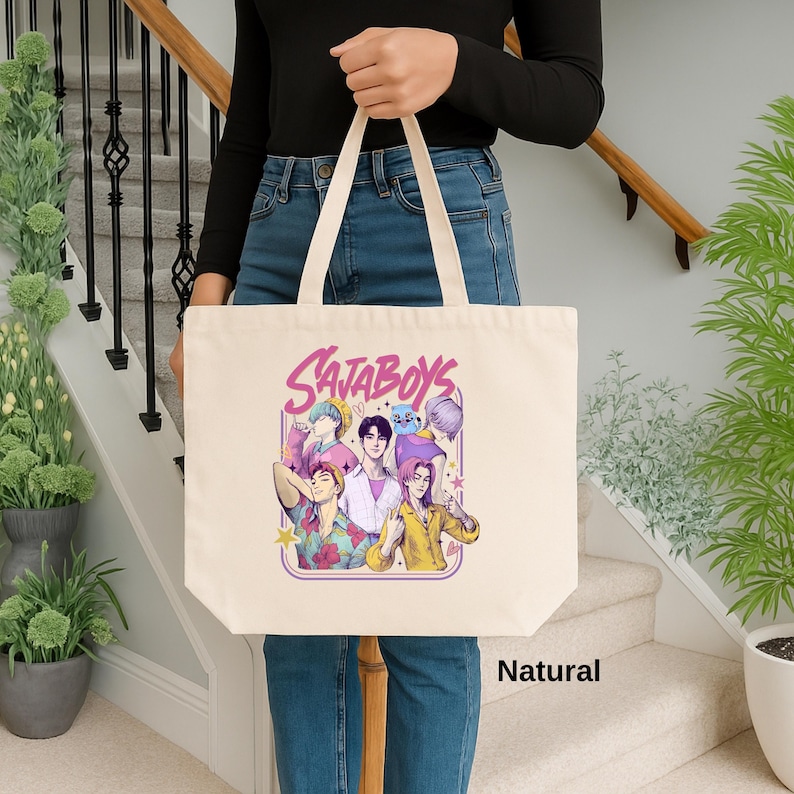 May include: A natural canvas tote bag featuring a colorful graphic of a group of cartoon characters with the text "SAJABOYS" in a purple and pink font. The bag is held by a person in blue jeans and a black top.