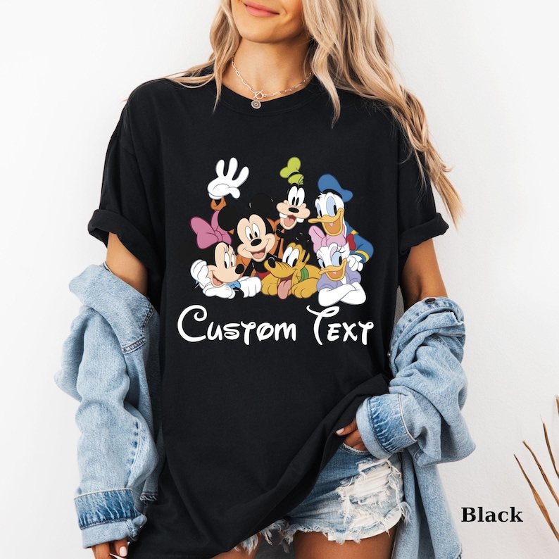 May include: Black t-shirt with a colourful graphic of Disney characters, including Mickey and Minnie Mouse, Goofy, Donald and Daisy Duck. The words "Custom Text" are written in white script. The shirt is a casual style.