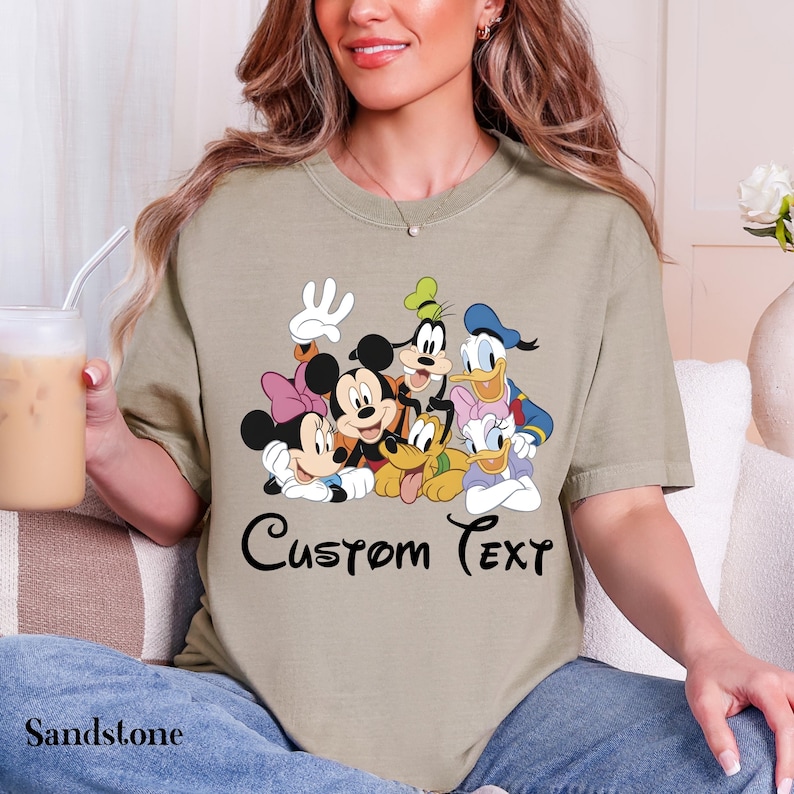 May include: Sandstone-coloured t-shirt with a graphic of Disney characters: Mickey Mouse, Minnie Mouse, Donald Duck, Daisy Duck, Goofy, and Pluto. The words "Custom Text" are written in black script. The shirt has a relaxed fit.