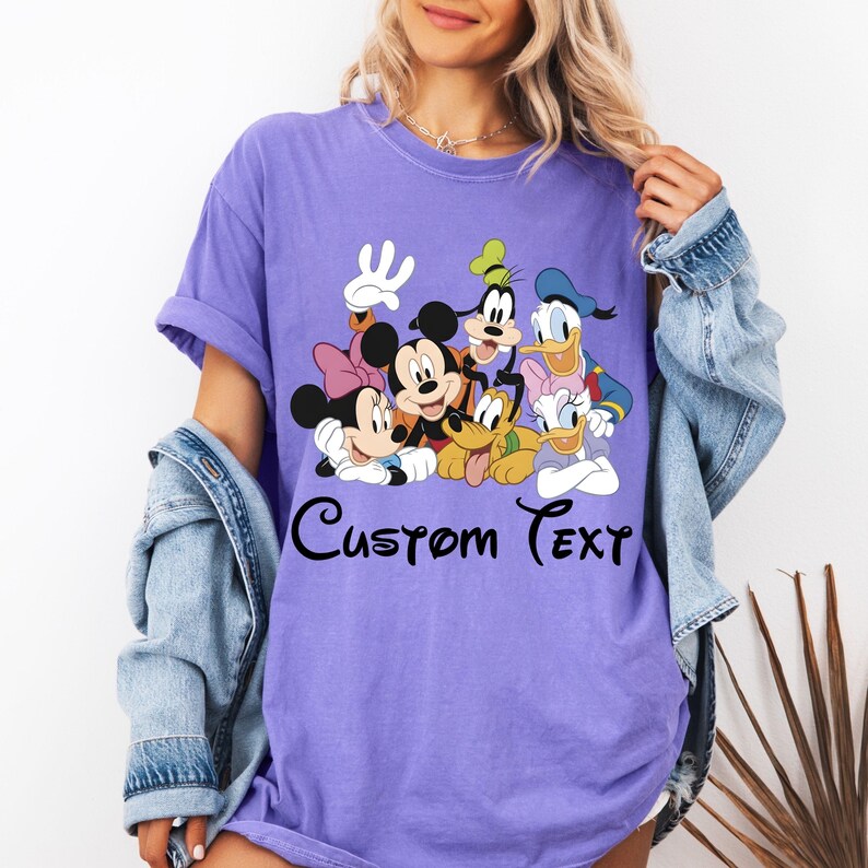 May include: A lilac t-shirt with a graphic of Disney characters, including Mickey Mouse, Minnie Mouse, Donald Duck, Daisy Duck, Goofy, and Pluto. The text "Custom Text" is written in a script font below the characters.