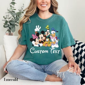 May include: Emerald green t-shirt with a cartoon graphic of Disney characters, including Mickey and Minnie Mouse, Donald Duck, and Goofy. The words "Custom Text" are written in white script. The word "Emerald" is printed at the bottom.