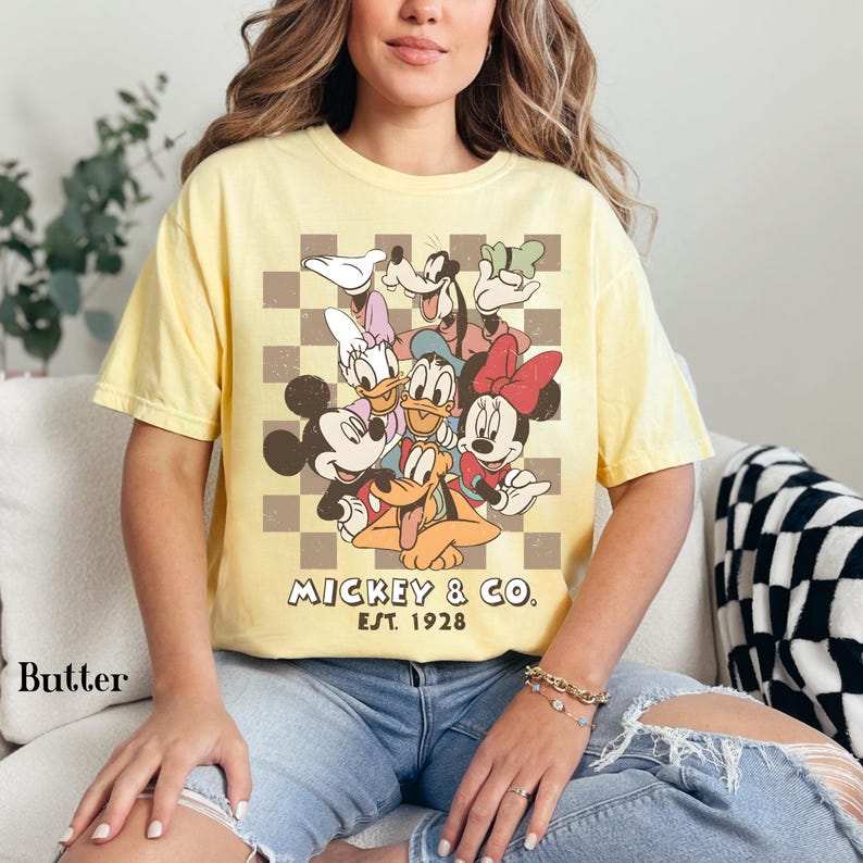May include: Butter yellow t-shirt featuring a graphic of Mickey Mouse and friends. The design includes Mickey, Minnie, Donald, Daisy, Goofy, and Pluto, set against a checkered background. The text "Mickey & Co. Est. 1928" is also visible.