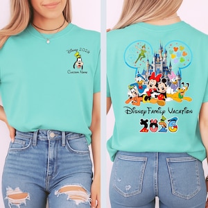 Op de afbeelding: Een lichtblauwgroen T-shirt met Disney-personages. De voorkant heeft een Goofy-afbeelding en de tekst "Disney 2026" en "Custom Name". De achterkant heeft een Disney-kasteelafbeelding, de tekst "Disney Family Vacation 2026" en andere Disney-personages.
