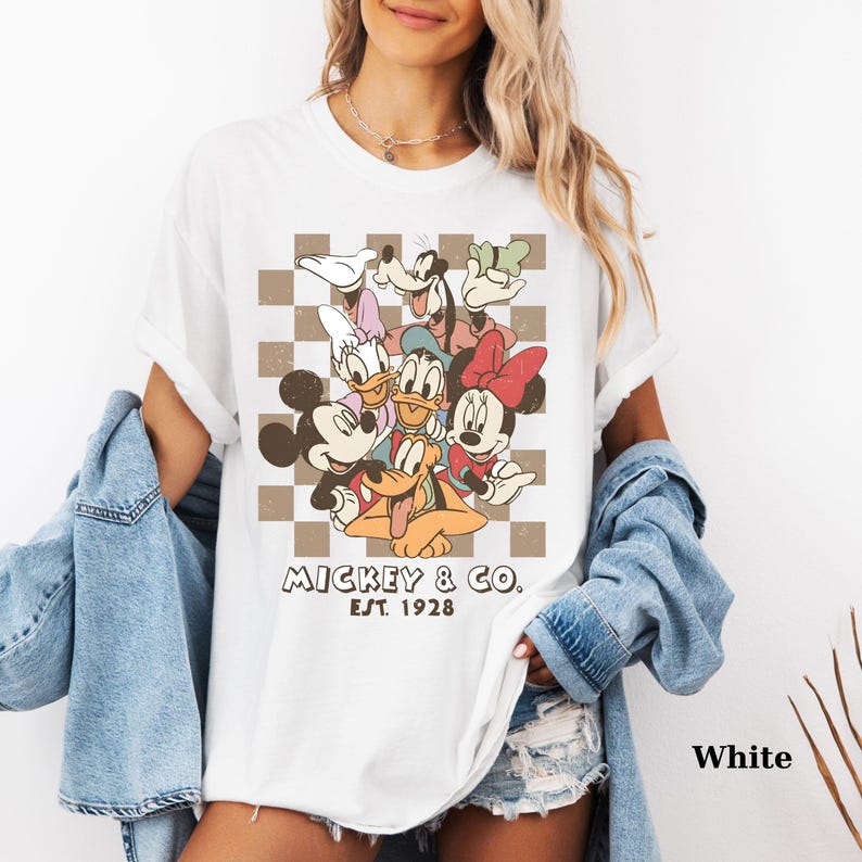 May include: White t-shirt featuring a graphic of Mickey Mouse, Minnie Mouse, Donald Duck, Daisy Duck, Goofy, and Pluto. The design is set against a brown and white checkered background, with "Mickey & Co. Est. 1928" printed below.