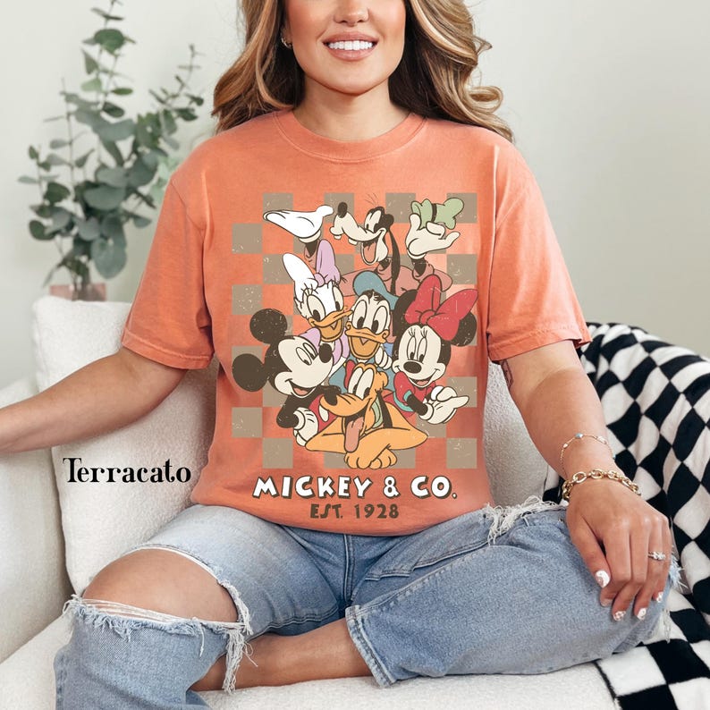 May include: Terracotta-colored t-shirt featuring a graphic of Mickey Mouse and friends. The design includes Mickey, Minnie, Donald, Daisy, Goofy, and Pluto, set against a checkered background. The text "Mickey & Co. Est. 1928" is printed below the characters.