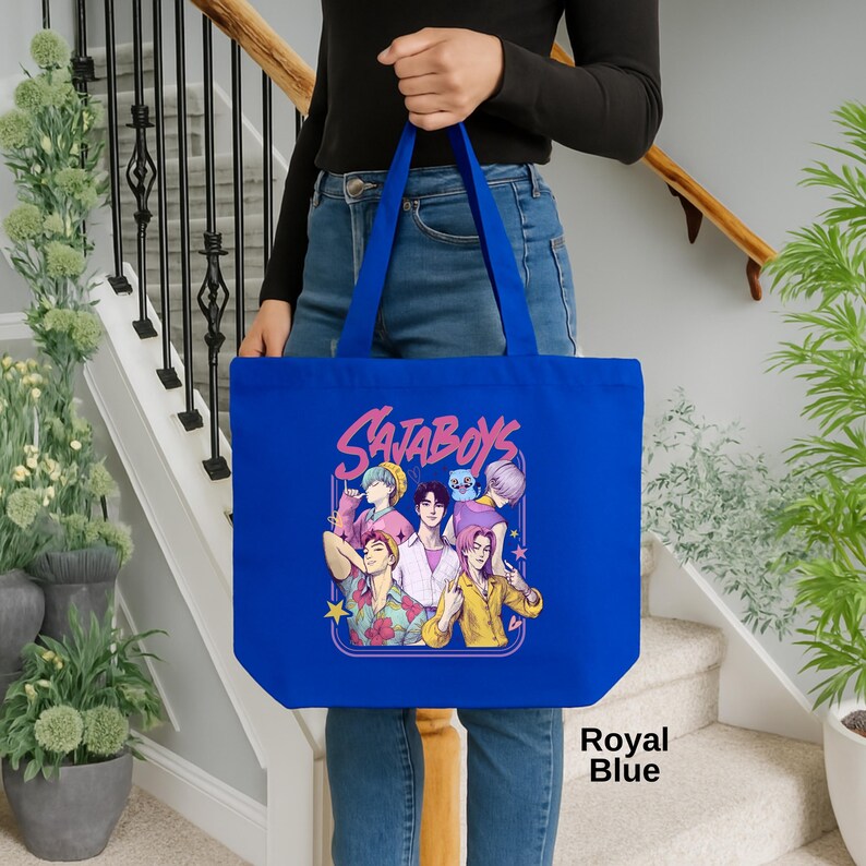 May include: Royal blue tote bag featuring the text "Sajaboys" and a colorful graphic of six cartoon figures. The bag has long handles and is held by a person. The bag is made of a sturdy material.