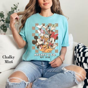 May include: A light blue t-shirt featuring a graphic of Mickey & Co. characters on a brown and white checkered background. The text "Mickey & Co. Est. 1928" is printed below the characters. The shirt is labeled "Chalky Mint".