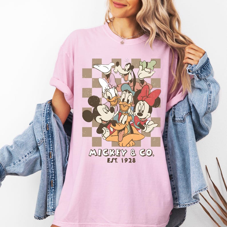 May include: A light pink t-shirt featuring a graphic of Mickey Mouse and friends against a brown and white checkered background. The text "Mickey & Co. Est. 1928" is printed below the characters. The shirt has short sleeves.