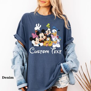 May include: A navy blue t-shirt with a graphic of Mickey Mouse, Minnie Mouse, Goofy, Pluto, Donald Duck, and Daisy Duck. The words "Custom Text" are written in white script below the characters. The word "Denim" is written in the bottom left corner.