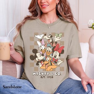 May include: Sandstone-colored t-shirt featuring a graphic of Mickey Mouse and friends. The design includes Mickey, Minnie, Donald, Daisy, Goofy, and Pluto, with the text "Mickey & Co. Est. 1928". The shirt has a classic, vintage aesthetic.