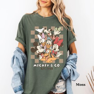 May include: Moss green t-shirt featuring a graphic of Mickey Mouse and friends, including Donald Duck and Minnie Mouse, on a checkered background. The text "Mickey & Co. EST 1928" is printed below the characters. The shirt is a relaxed fit.
