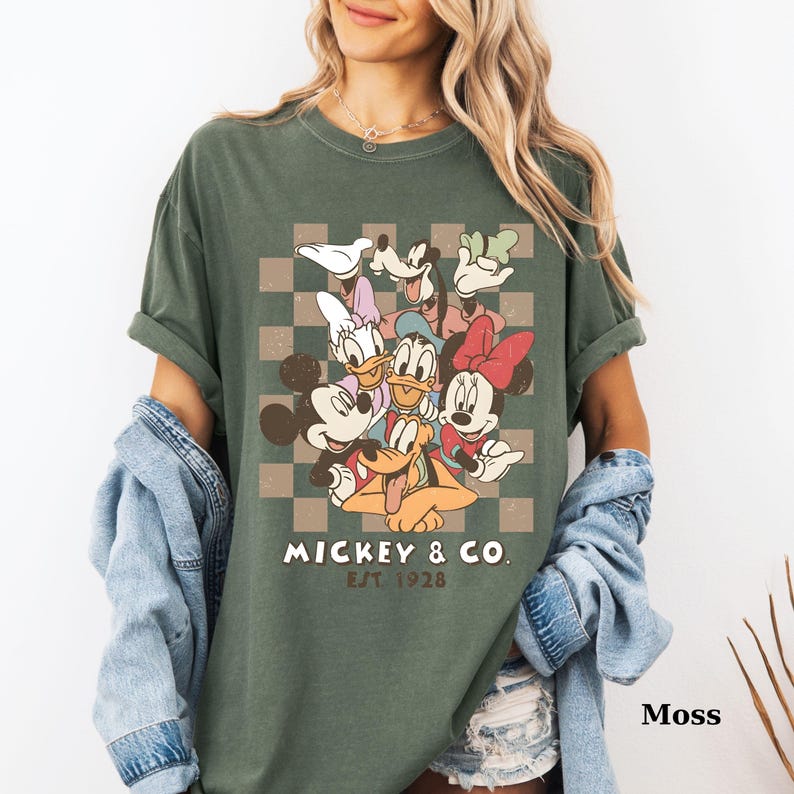 May include: Moss green t-shirt featuring a graphic of Mickey Mouse and friends, including Donald Duck and Minnie Mouse, on a checkered background. The text "Mickey & Co. EST 1928" is printed below the characters. The shirt is a relaxed fit.
