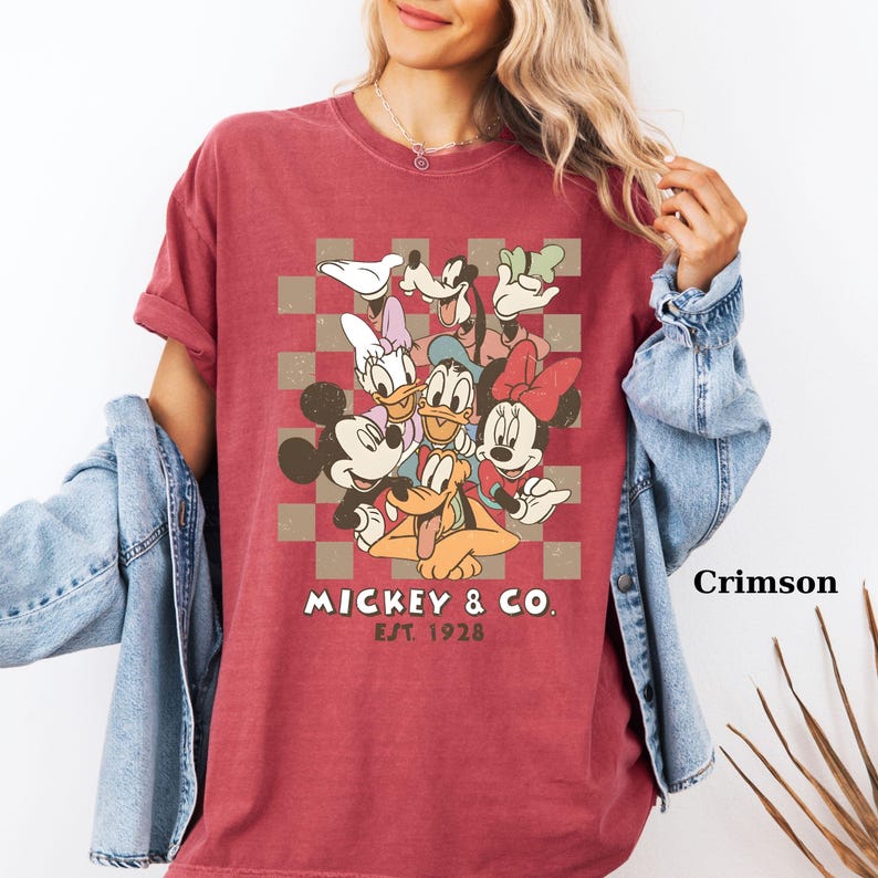 May include: Crimson-colored t-shirt featuring a graphic of Mickey Mouse and friends. The design includes Mickey, Minnie, Donald, Daisy, Goofy, and Pluto, set against a checkered background. The text "Mickey & Co. Est. 1928" is printed below the characters.
