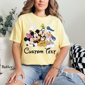 May include: A pale yellow t-shirt with a graphic of Disney characters such as Mickey Mouse, Minnie Mouse, Goofy, Pluto, Donald Duck, and Daisy Duck. The words "Custom Text" are written in a script font. The shirt is a butter colour.