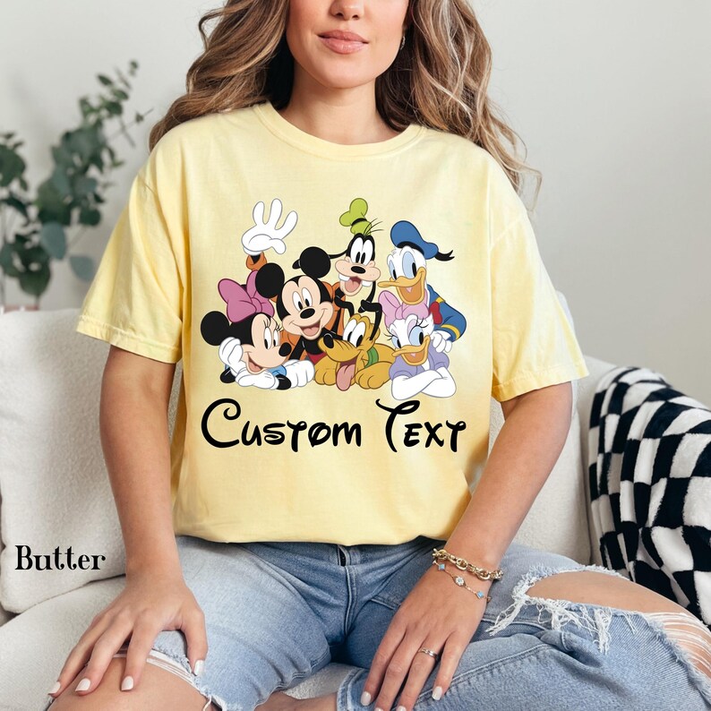 May include: A pale yellow t-shirt with a graphic of Disney characters such as Mickey Mouse, Minnie Mouse, Goofy, Pluto, Donald Duck, and Daisy Duck. The words "Custom Text" are written in a script font. The shirt is a butter colour.