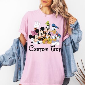 May include: A pale pink t-shirt with a cartoon graphic of Mickey Mouse, Minnie Mouse, Goofy, Donald Duck, Daisy Duck, and Pluto. The words "Custom Text" are written in black script below the characters. The shirt has a relaxed fit.