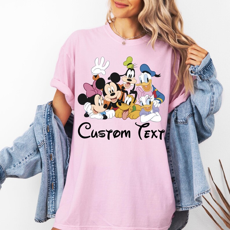 May include: A pale pink t-shirt with a cartoon graphic of Mickey Mouse, Minnie Mouse, Goofy, Donald Duck, Daisy Duck, and Pluto. The words "Custom Text" are written in black script below the characters. The shirt has a relaxed fit.