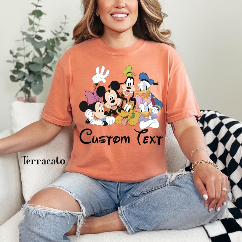 May include: A terracotta-coloured t-shirt displaying a group of cartoon characters, including Mickey Mouse, Minnie Mouse, Goofy, Donald Duck, and Daisy Duck. The words "Custom Text" are written in a script font below the characters. The shirt is worn by a person.
