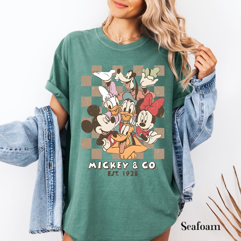 May include: Seafoam green t-shirt featuring a graphic of Mickey Mouse and friends on a checkered background. The design includes Mickey, Minnie, Donald, Daisy, Goofy, and Pluto. The text "Mickey & Co. Est. 1928" is printed below the characters.