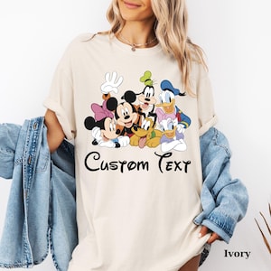 May include: An ivory-coloured t-shirt with a cartoon graphic of Mickey Mouse, Minnie Mouse, Donald Duck, and Goofy. The words "Custom Text" are written in black script. The shirt is a loose fit and the colour is described as Ivory.