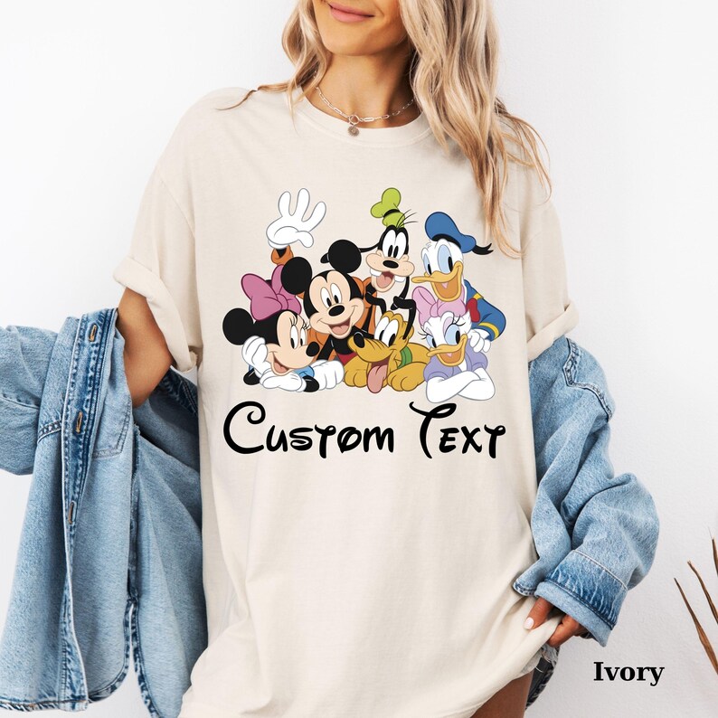 May include: An ivory-coloured t-shirt with a cartoon graphic of Mickey Mouse, Minnie Mouse, Donald Duck, and Goofy. The words "Custom Text" are written in black script. The shirt is a loose fit and the colour is described as Ivory.