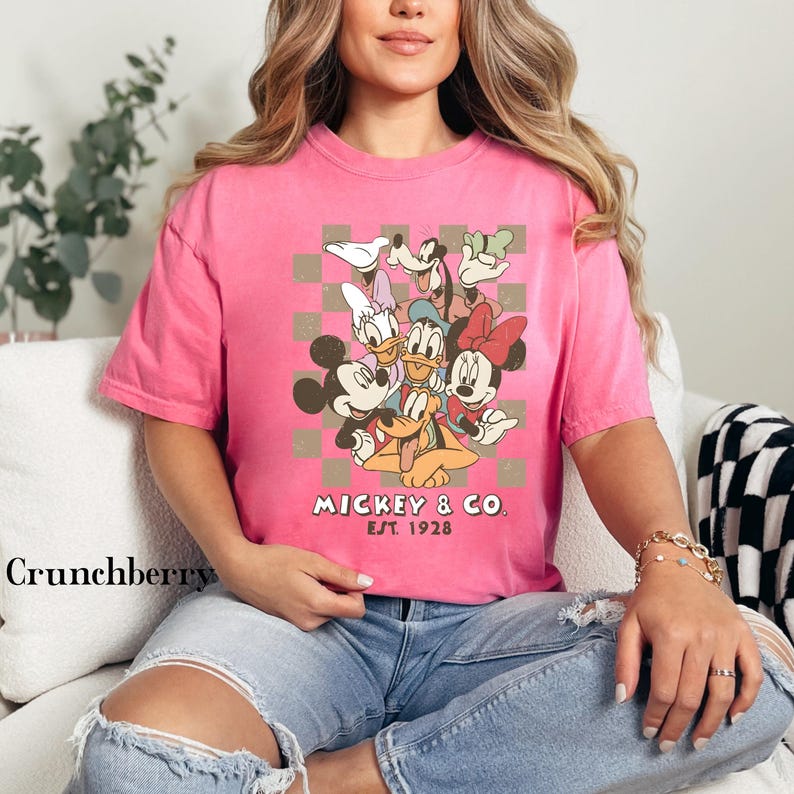 May include: A pink t-shirt featuring a graphic of Mickey Mouse and friends, including Minnie Mouse, Donald Duck, and Goofy, set against a checkered background. The text "Mickey & Co. Est. 1928" is printed below the characters.