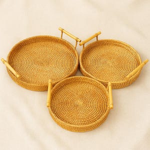 May include: Three round woven rattan trays with bamboo handles. The trays are a light brown color and have a circular woven pattern. The handles are attached to the sides of the trays. The trays are of varying sizes.