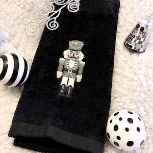 May include: Black hand towel featuring an embroidered nutcracker design in grey, white, and black. The towel is folded and displayed with festive decorations, including a black and white striped bauble and a white polka dot bauble.