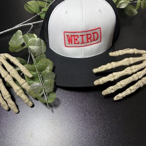 Può includere: Cappellino da camionista bianco e nero con la scritta "WEIRD" ricamata in rosso sul davanti. Il cappellino ha un pannello frontale bianco e un retro in rete nera. Il testo è all'interno di un rettangolo rosso.