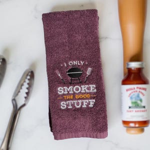 May include: A purple kitchen towel with the text "I only smoke the good stuff" embroidered on it. The text is in white with orange accents. A grill icon is above the text.