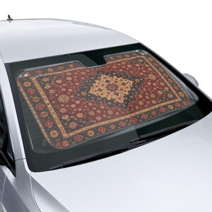 May include: A car windshield sunshade featuring a detailed oriental rug design. The sunshade displays a central medallion in beige and gold, surrounded by a deep red field with floral patterns and a navy blue border. The sunshade is installed in a silver car.
