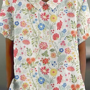 Può includere: Camicia bianca a maniche corte con un motivo floreale colorato. Il design presenta fiori rossi, gialli, blu e rosa con foglie e steli verdi. La camicia ha un colletto rotondo.