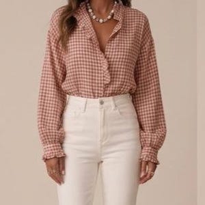Könnte beinhalten: Eine Frau trägt ein langärmliges, geknöpftes Hemd mit rosa-weißem Vichy-Muster und eine weiße Jeans. Das Hemd hat eine Rüschenverzierung und die Jeans sind hell.