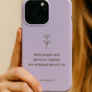 Peut inclure: Coque de téléphone lilas avec un motif floral et le texte "La force et la dignité sont ses vêtements" de Proverbes 31:25. L'étui a une découpe pour l'appareil photo et est tenu par une personne.