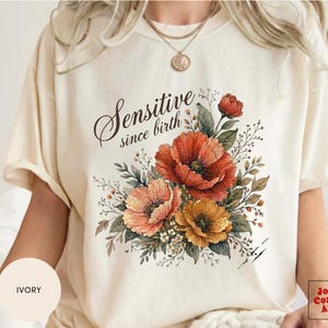 Puede incluir: Una camiseta de manga corta color marfil con un estampado gráfico de un ramo de flores con amapolas rojas, naranjas y amarillas y follaje verde. El texto "Sensitive since birth" está impreso en una letra cursiva negra sobre las flores.