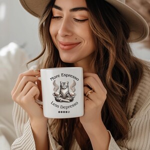 Puede incluir: Taza de cerámica blanca con una ilustración en blanco y negro de un gato meditando con dos tazas de café. El texto en la taza dice "More Espresso Less Depresso".