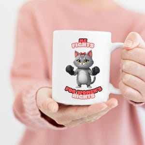 Puede incluir: Taza de cerámica blanca con un gato gris de dibujos animados que lleva guantes de boxeo y un lazo rosa. La taza tiene el texto "ME FIGHTS FOR WOMEN'S RIGHTS" en rosa y negro.