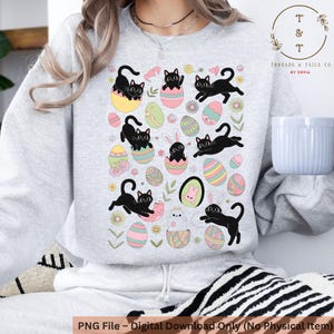 May include: A grey sweatshirt featuring a black cat and Easter egg design. The design depicts black cats emerging from colourful Easter eggs, some with pink bunny ears. The design is surrounded by flowers and leaves.