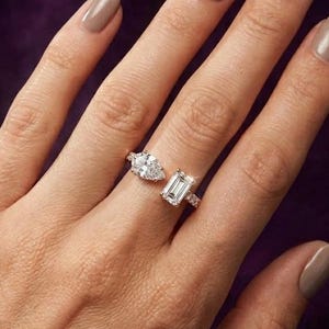 Może przedstawiać: Dwa diamentowe pierścionki na dłoni z neutralnym lakierem do paznokci. Jeden pierścionek ma diament w kształcie gruszki, drugi diament o szlifie szmaragdowym. Oba osadzone w złotej oprawie.