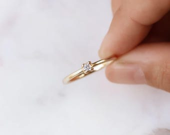 May include: A delicate gold ring with a small, square-cut diamond set in the center. The ring is held between fingers against a soft, blurred background. The band is thin and polished, reflecting light.