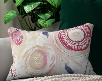 Può includere: Cuscino lombare decorativo con un design ad acquerello nei toni del rosa, beige e bianco. Il cuscino presenta un motivo circolare ed elementi floreali astratti. È posto su un divano grigio con una coperta color crema.