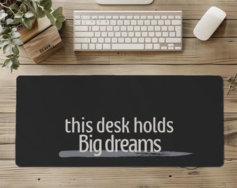 May include: A black desk mat with the text "this desk holds Big dreams" in white. A grey brushstroke underlines the text. The mat is on a wooden desk with a keyboard, mouse, and a small plant.