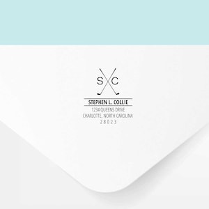 May include: A white envelope with a black return address label. The label features a logo of two crossed golf clubs with the letters "SXC" above them. The text below the logo reads "Stephen L. Collie, 1234 Queens Drive, Charlotte, North Carolina 28023".