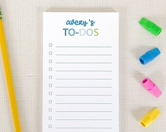 May include: A white notepad with a blue and green gradient design. The notepad has the text "avery's TO-DOS" written on the top. There are 14 lines on the notepad for writing. A yellow pencil is on the left side of the notepad. There are four colorful pencil toppers on the right side of the notepad.